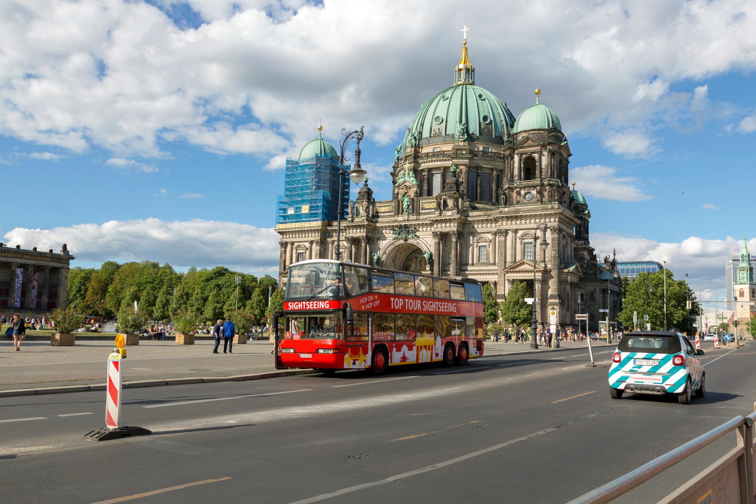 Double-decker sightseeing bus passes Berlin Cathedral under a bright blue sky.